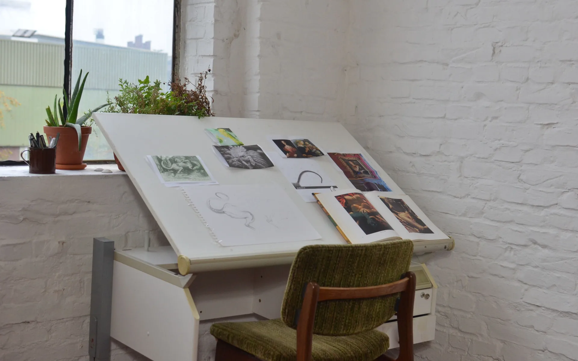 Une table à dessin inclinée près d'une fenêtre : reproductions de peintures, carnet à spirale avec un croquis au crayon et livre d'art ouvert, devant une chaise d'atelier verte