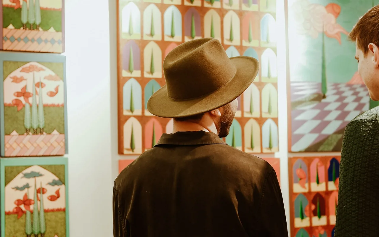 Deux visiteurs dans une galerie : un homme au chapeau vu de dos et, à droite, un homme en pull vert, devant des tableaux aux arcades colorées accrochés au mur blanc