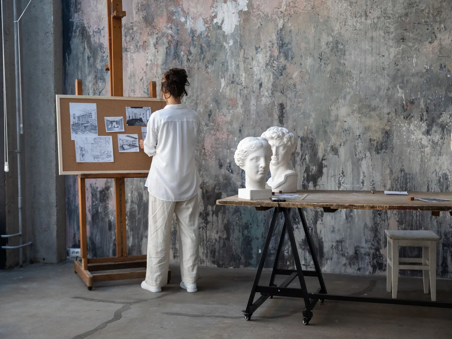 Une artiste de dos épingle des dessins sur un panneau, à côté de bustes en plâtre, dans un atelier aux murs patinés