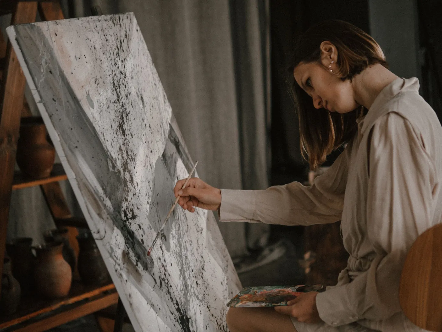 Une artiste en chemise de lin peint une grande toile abstraite grise et blanche posée sur un chevalet