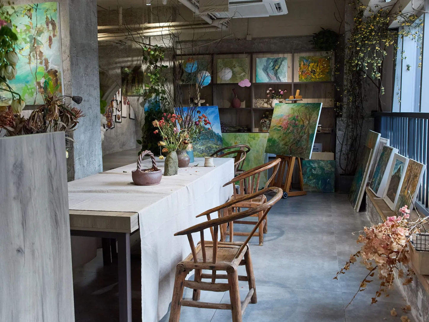 Un atelier-galerie clair : une longue table nappée de lin, des chaises en bois, des fleurs séchées et des toiles colorées le long des murs