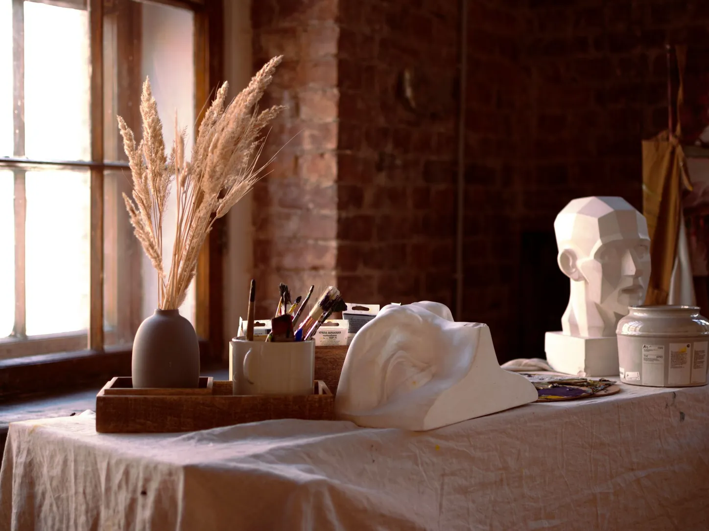 Une table d'atelier nappée de lin près d'une fenêtre : têtes en plâtre, pinceaux en pot et bouquet d'herbes sèches devant un mur de briques