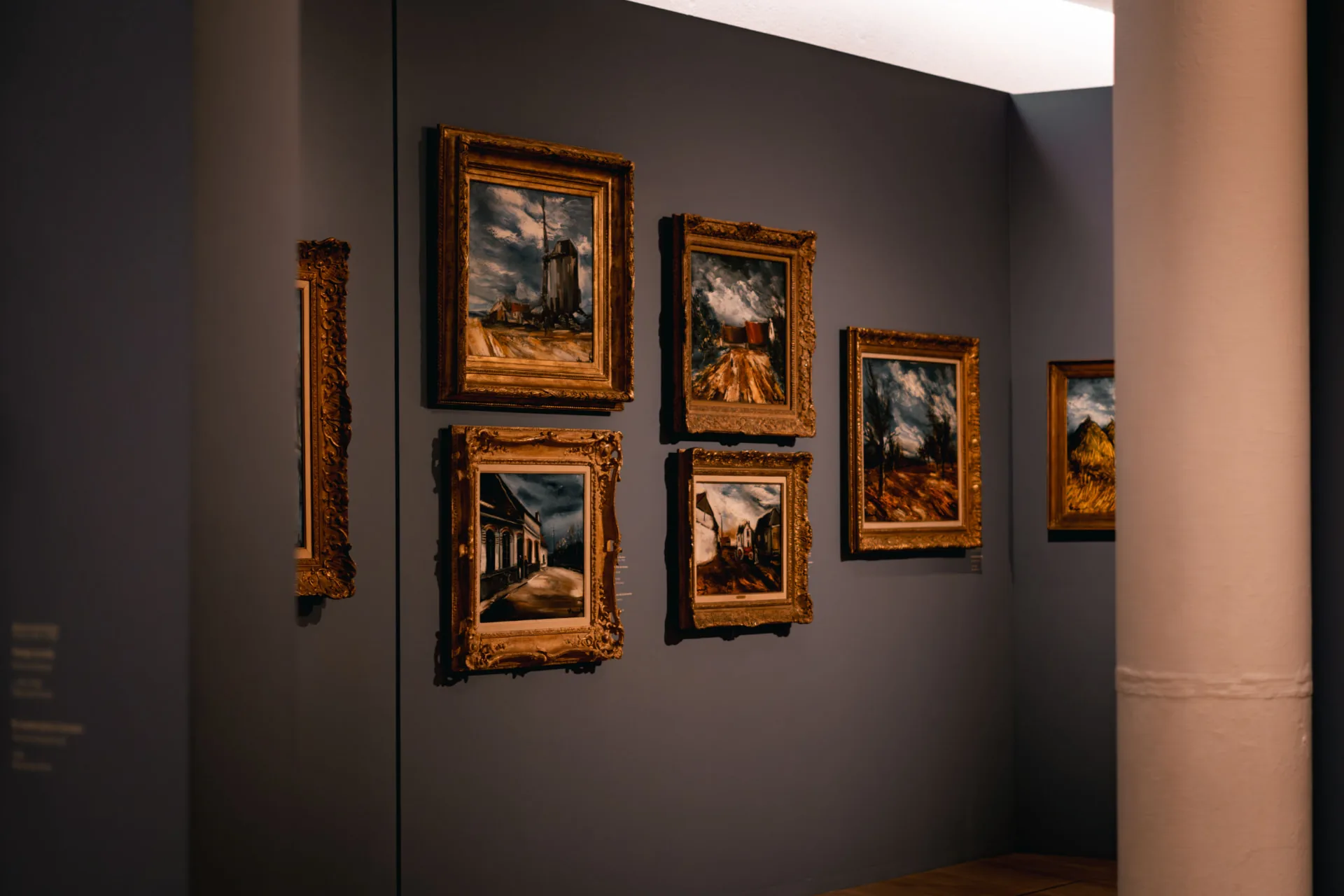 Une salle de musée aux murs gris : tableaux de paysages dans de larges cadres dorés, colonne blanche à droite, sous une lumière chaude