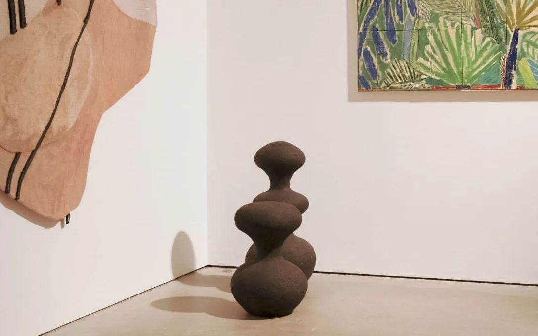 Une galerie aux murs clairs : grande tapisserie abstraite accrochée à gauche, sculpture sombre posée au sol au centre, tableau de feuillages tropicaux à droite